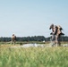 Two military personnel in desert camouflage uniforms are gathering parachutes in an open grassy field after a landing operation. The foreground soldier is actively collecting parachute lines and canopy material, while a second soldier is visible in the background doing the same. The setting appears to be a designated drop zone with a tree line visible on the horizon.