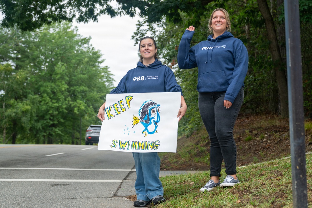 Two young women stand on the side of a road wearing matching navy blue '988 Suicide &amp; Crisis Lifeline' hoodies. One holds a hand-drawn sign reading 'Keep Swimming' featuring a cartoon fish resembling Dory from Finding Nemo. They appear to be cheering on participants in a race or walk event.