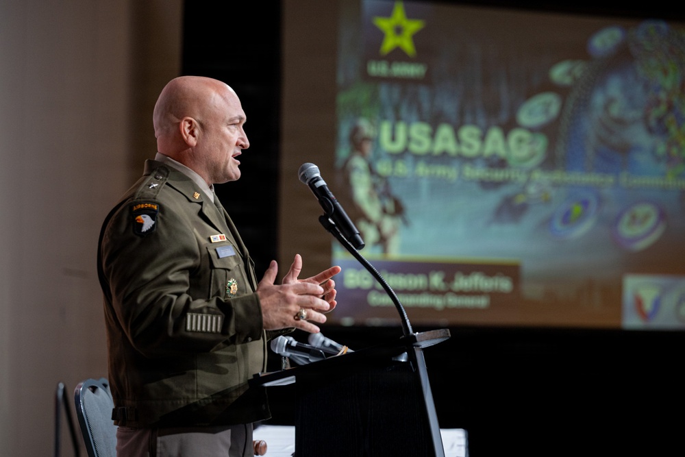 A U.S. Army officer in dress uniform is delivering a speech at a podium with a microphone. The presentation slide behind him displays 'USASAC' (U.S. Army Security Assistance Command) along with a name placard partially reading 'BG...Jason K. Jeffers, Commanding General.' The setting appears to be a formal military conference or briefing event.