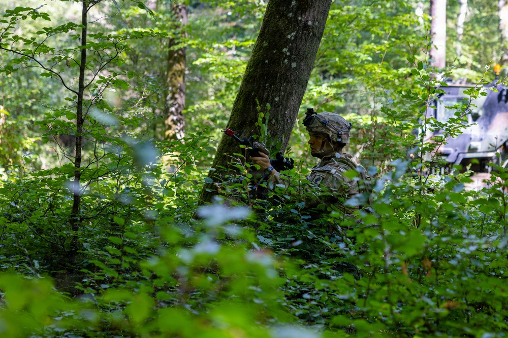 A soldier in full combat gear is positioned behind a large tree trunk in a dense forest, holding a rifle in a tactical stance. The soldier is wearing a camouflage uniform, helmet with mounted equipment, and appears to be conducting a military exercise or patrol. A military armored vehicle is partially visible in the background to the right.