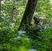 A soldier in full combat gear is positioned behind a large tree trunk in a dense forest, holding a rifle in a tactical stance. The soldier is wearing a camouflage uniform, helmet with mounted equipment, and appears to be conducting a military exercise or patrol. A military armored vehicle is partially visible in the background to the right.