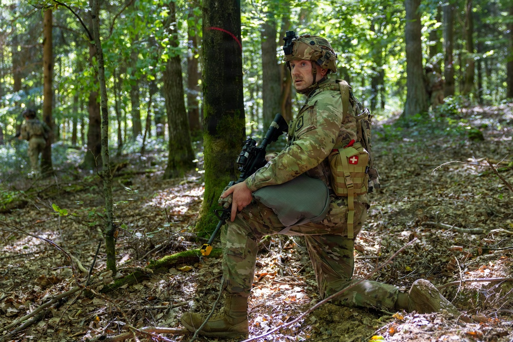 A soldier in full combat gear is kneeling beside a tree in a forested environment, appearing alert and scanning his surroundings. He is equipped with a rifle, tactical vest, helmet with night vision mount, and a medical pouch marked with a red cross. At least two additional soldiers are visible in the background, suggesting a patrol or training exercise.