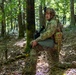A soldier in full combat gear is kneeling beside a tree in a forested environment, appearing alert and scanning his surroundings. He is equipped with a rifle, tactical vest, helmet with night vision mount, and a medical pouch marked with a red cross. At least two additional soldiers are visible in the background, suggesting a patrol or training exercise.