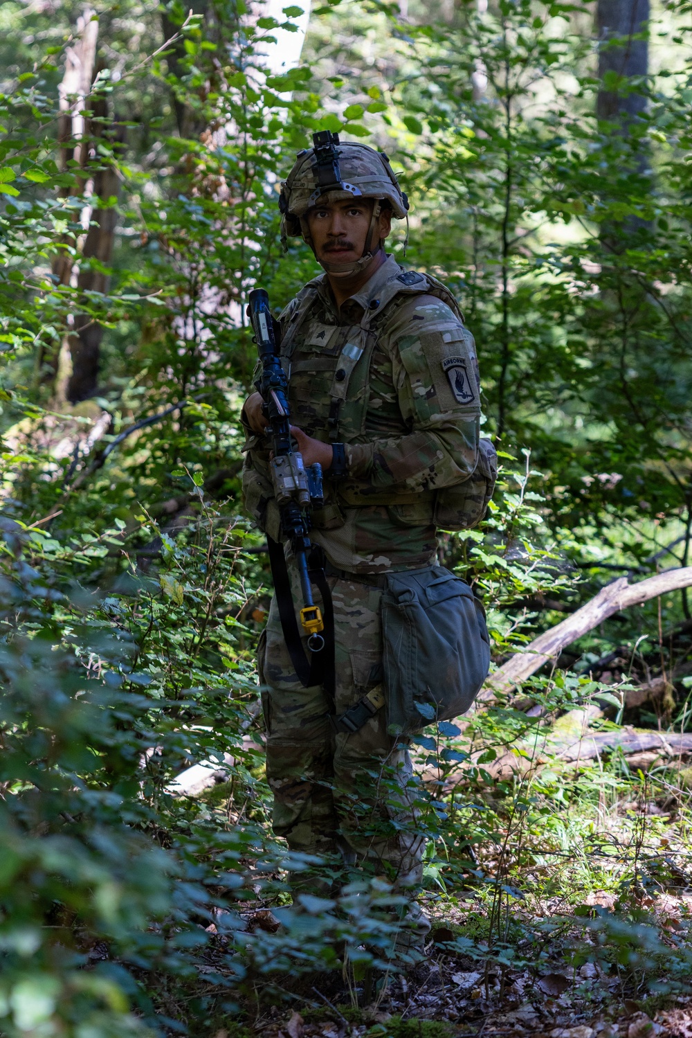 A U.S. Army soldier in full combat gear stands alert in a dense woodland environment. The soldier is wearing Operational Camouflage Pattern (OCP) uniform with an Airborne patch visible on the left shoulder, carrying a rifle equipped with training accessories. The setting appears to be a field training exercise based on the MILES (Multiple Integrated Laser Engagement System) gear attached to the weapon.