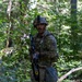 A U.S. Army soldier in full combat gear stands alert in a dense woodland environment. The soldier is wearing Operational Camouflage Pattern (OCP) uniform with an Airborne patch visible on the left shoulder, carrying a rifle equipped with training accessories. The setting appears to be a field training exercise based on the MILES (Multiple Integrated Laser Engagement System) gear attached to the weapon.