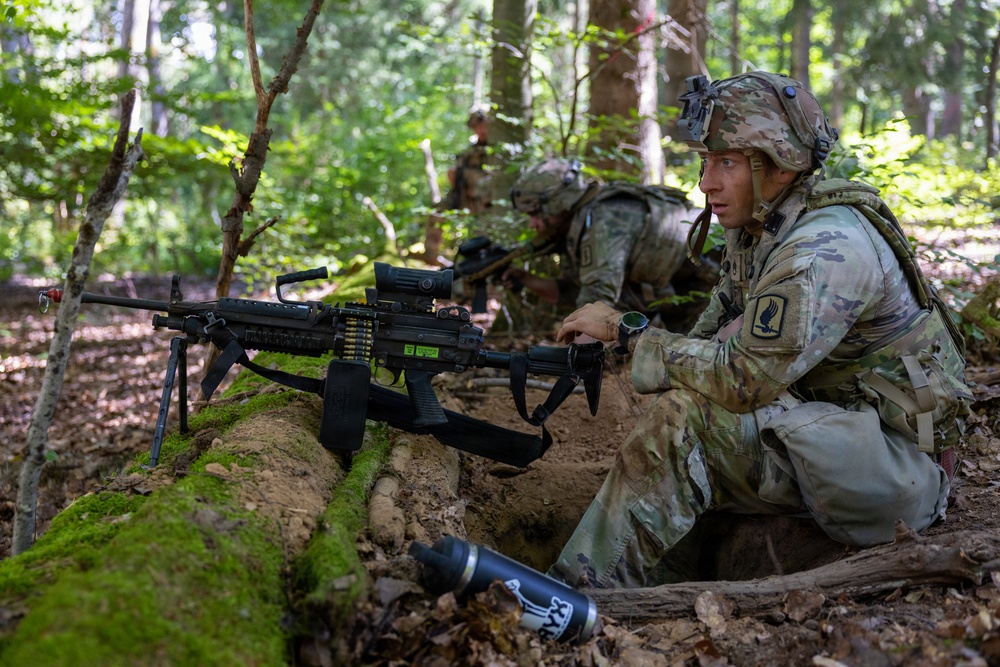 Military personnel in camouflage uniforms are positioned in a forested area behind a moss-covered fallen log, appearing to be conducting a tactical field exercise or training operation. The foreground soldier is equipped with a light machine gun mounted on a bipod with an ammunition belt loaded. Additional soldiers are visible in the background in similar tactical positions.