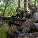 Military personnel in camouflage uniforms are positioned in a forested area behind a moss-covered fallen log, appearing to be conducting a tactical field exercise or training operation. The foreground soldier is equipped with a light machine gun mounted on a bipod with an ammunition belt loaded. Additional soldiers are visible in the background in similar tactical positions.