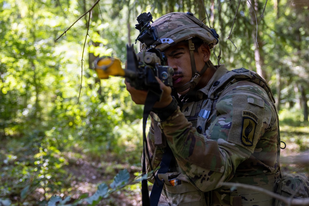 A U.S. Army Airborne soldier in full combat gear is aiming a rifle with a yellow training/blank-firing attachment in a forested environment. The soldier wears MultiCam (OCP) camouflage, a tactical helmet with night-vision mount, and a plate carrier vest. The setting and equipment suggest a training exercise rather than live combat.