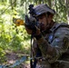 A U.S. Army Airborne soldier in full combat gear is aiming a rifle with a yellow training/blank-firing attachment in a forested environment. The soldier wears MultiCam (OCP) camouflage, a tactical helmet with night-vision mount, and a plate carrier vest. The setting and equipment suggest a training exercise rather than live combat.