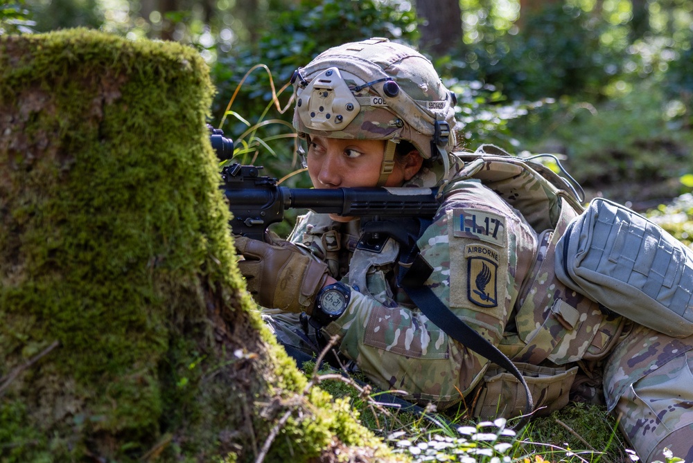 A U.S. Army soldier from the 17th Airborne division is positioned prone behind a moss-covered tree in a forested environment, aiming a rifle with an optical sight. The soldier is wearing full MultiCam camouflage gear including a tactical helmet with accessories and a tactical backpack. The scene appears to depict a field training exercise or tactical operation in a woodland setting.