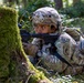 A U.S. Army soldier from the 17th Airborne division is positioned prone behind a moss-covered tree in a forested environment, aiming a rifle with an optical sight. The soldier is wearing full MultiCam camouflage gear including a tactical helmet with accessories and a tactical backpack. The scene appears to depict a field training exercise or tactical operation in a woodland setting.