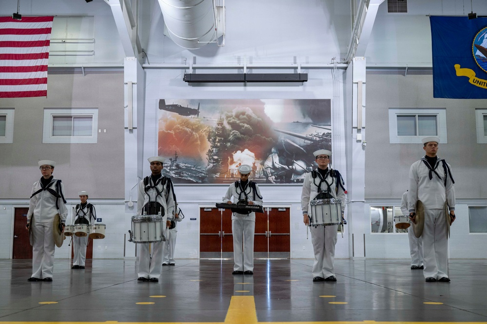 A group of U.S. Navy sailors in dress white uniforms are positioned in formation inside a large indoor facility, appearing to be a military band or drum line. They are holding various percussion instruments including snare drums and a keyboard/synthesizer. Behind them is a large projected image depicting what appears to be a naval battle scene with fire and explosions, likely referencing Pearl Harbor or a similar historical naval engagement.