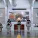 A group of U.S. Navy sailors in dress white uniforms are positioned in formation inside a large indoor facility, appearing to be a military band or drum line. They are holding various percussion instruments including snare drums and a keyboard/synthesizer. Behind them is a large projected image depicting what appears to be a naval battle scene with fire and explosions, likely referencing Pearl Harbor or a similar historical naval engagement.