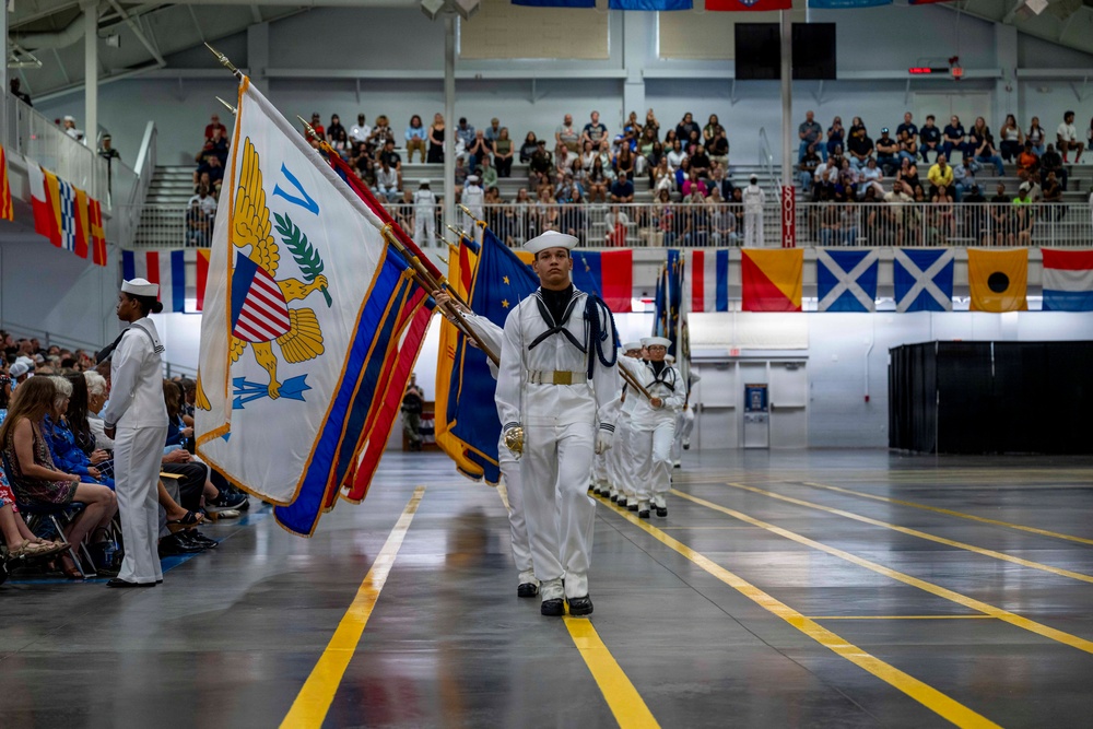 A U.S. Navy color guard ceremony is taking place inside a large indoor arena or drill hall. Sailors in white dress uniforms are marching in formation while carrying multiple flags, including what appears to be the Secretary of the Navy flag and other military colors. A large audience is seated in bleachers and along the floor level observing the formal ceremony.