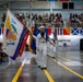 A U.S. Navy color guard ceremony is taking place inside a large indoor arena or drill hall. Sailors in white dress uniforms are marching in formation while carrying multiple flags, including what appears to be the Secretary of the Navy flag and other military colors. A large audience is seated in bleachers and along the floor level observing the formal ceremony.