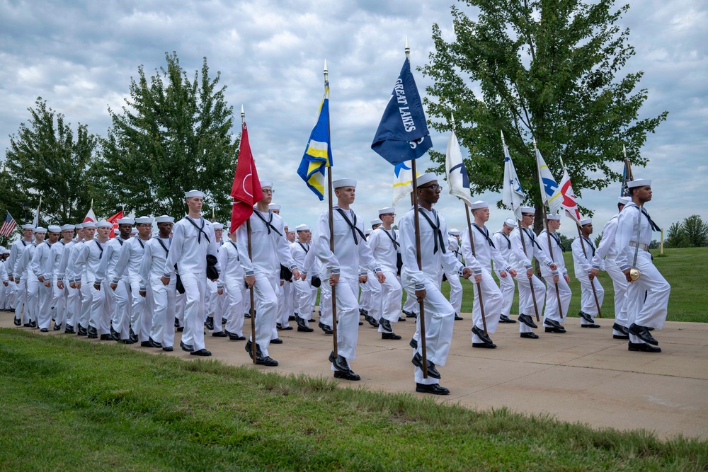 A large formation of U.S. Navy recruits or sailors dressed in white dress uniforms are marching in a parade or graduation ceremony. Multiple color guard members carry unit guidons and flags, including one clearly marked 'Great Lakes,' indicating this is likely a recruit training graduation at Naval Station Great Lakes, Illinois. The formation is well-organized and marching in step along a paved pathway surrounded by green grass and trees.