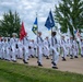 A large formation of U.S. Navy recruits or sailors dressed in white dress uniforms are marching in a parade or graduation ceremony. Multiple color guard members carry unit guidons and flags, including one clearly marked 'Great Lakes,' indicating this is likely a recruit training graduation at Naval Station Great Lakes, Illinois. The formation is well-organized and marching in step along a paved pathway surrounded by green grass and trees.