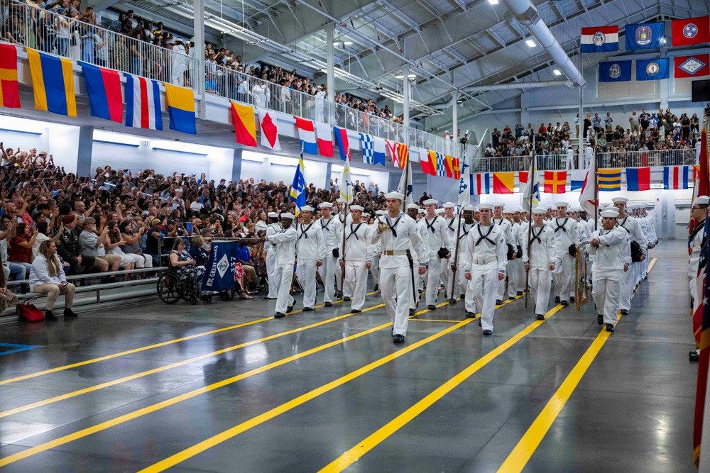 A U.S. Navy recruit graduation ceremony (Pass in Review) is taking place inside a large indoor drill hall, likely at Recruit Training Command (RTC) Great Lakes, Illinois. Rows of recruits dressed in white dress uniforms march in formation while carrying flags and guidons before a large crowd of spectators filling the bleachers and balcony. The hall is decorated with numerous nautical signal flags and U.S. state flags hanging from the ceiling and walls.