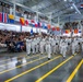A U.S. Navy recruit graduation ceremony (Pass in Review) is taking place inside a large indoor drill hall, likely at Recruit Training Command (RTC) Great Lakes, Illinois. Rows of recruits dressed in white dress uniforms march in formation while carrying flags and guidons before a large crowd of spectators filling the bleachers and balcony. The hall is decorated with numerous nautical signal flags and U.S. state flags hanging from the ceiling and walls.