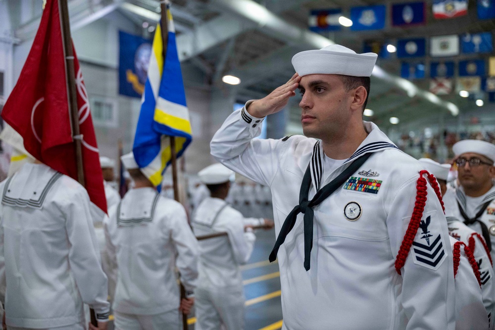 A U.S. Navy sailor in dress white uniform renders a salute during a formal military ceremony held indoors, likely at a naval training or ceremonial facility. The sailor in the foreground displays multiple service ribbons, a Petty Officer rating badge, and red aiguillette cords, indicating a senior enlisted role. Multiple sailors and color guard members with flags are visible in the background.