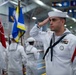 A U.S. Navy sailor in dress white uniform renders a salute during a formal military ceremony held indoors, likely at a naval training or ceremonial facility. The sailor in the foreground displays multiple service ribbons, a Petty Officer rating badge, and red aiguillette cords, indicating a senior enlisted role. Multiple sailors and color guard members with flags are visible in the background.