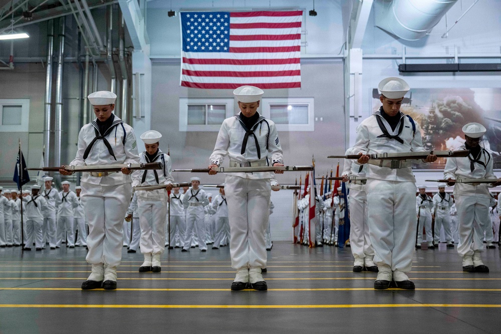 A group of U.S. Navy personnel dressed in white dress uniforms are performing a ceremonial rifle drill inside a large indoor facility. A large American flag is prominently displayed on the wall in the background, and rows of additional sailors in formation are visible behind the drill team. The scene appears to be a formal military graduation or pass-in-review ceremony.