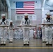 A group of U.S. Navy personnel dressed in white dress uniforms are performing a ceremonial rifle drill inside a large indoor facility. A large American flag is prominently displayed on the wall in the background, and rows of additional sailors in formation are visible behind the drill team. The scene appears to be a formal military graduation or pass-in-review ceremony.