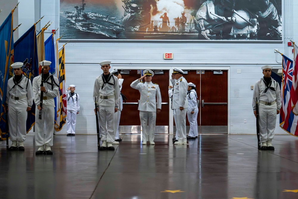 A formal U.S. Navy military ceremony is taking place inside an indoor venue, likely a naval base or training facility. Personnel in dress white uniforms are standing at attention and rendering salutes, with a color guard holding multiple flags including state and national flags. A large black-and-white naval mural depicting ships and combat scenes serves as a dramatic backdrop.