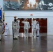 A formal U.S. Navy military ceremony is taking place inside an indoor venue, likely a naval base or training facility. Personnel in dress white uniforms are standing at attention and rendering salutes, with a color guard holding multiple flags including state and national flags. A large black-and-white naval mural depicting ships and combat scenes serves as a dramatic backdrop.