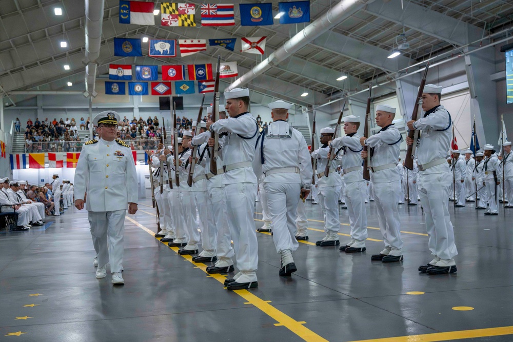 A U.S. Navy graduation or pass-in-review ceremony taking place inside a large indoor drill hall. Recruits in white dress uniforms are marching in formation while carrying rifles, being reviewed by a senior officer in dress whites with gold shoulder boards. State and territorial flags are displayed along the ceiling of the facility.