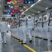 A U.S. Navy graduation or pass-in-review ceremony taking place inside a large indoor drill hall. Recruits in white dress uniforms are marching in formation while carrying rifles, being reviewed by a senior officer in dress whites with gold shoulder boards. State and territorial flags are displayed along the ceiling of the facility.