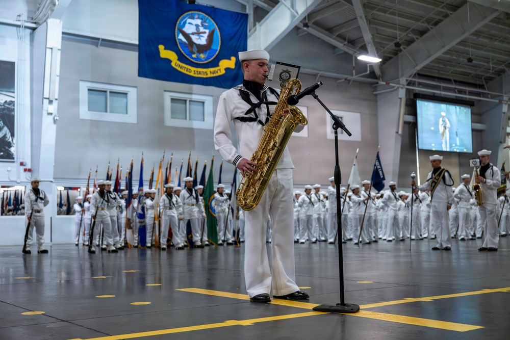 A United States Navy ceremonial event is taking place indoors, likely in a hangar bay or large military facility. A sailor in dress whites is performing a solo on a baritone saxophone in the foreground, while rows of sailors in formation and color guard members holding flags stand in the background. A large United States Navy flag is prominently displayed on the wall.