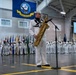 A United States Navy ceremonial event is taking place indoors, likely in a hangar bay or large military facility. A sailor in dress whites is performing a solo on a baritone saxophone in the foreground, while rows of sailors in formation and color guard members holding flags stand in the background. A large United States Navy flag is prominently displayed on the wall.