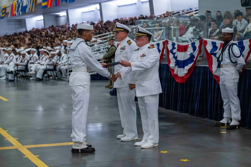 A U.S. Navy graduation ceremony is taking place in a large indoor venue, likely a training command facility. A senior naval officer is presenting a certificate or award to a graduating sailor in dress white uniform, while rows of graduates and a large audience of spectators look on. The setting is decorated with patriotic red, white, and blue bunting.