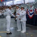 A U.S. Navy graduation ceremony is taking place in a large indoor venue, likely a training command facility. A senior naval officer is presenting a certificate or award to a graduating sailor in dress white uniform, while rows of graduates and a large audience of spectators look on. The setting is decorated with patriotic red, white, and blue bunting.