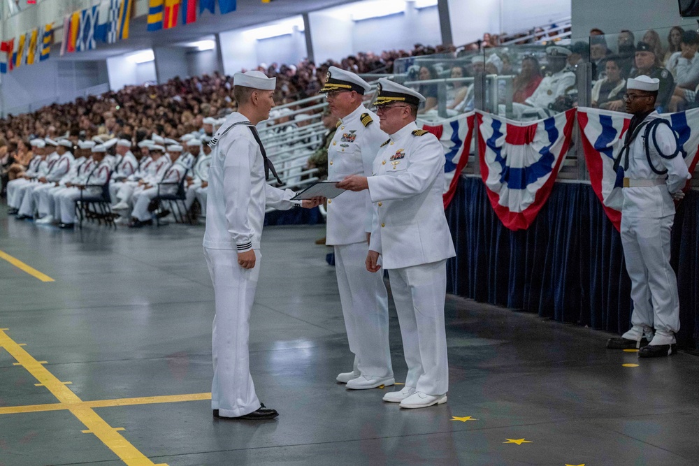 A formal U.S. Navy graduation or awards ceremony is taking place in a large indoor venue, likely a drill hall or gymnasium. A senior naval officer in dress whites is presenting a document or certificate to a junior enlisted sailor. The venue is filled with seated sailors in uniform and civilian spectators in the stands.