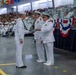 A formal U.S. Navy graduation or awards ceremony is taking place in a large indoor venue, likely a drill hall or gymnasium. A senior naval officer in dress whites is presenting a document or certificate to a junior enlisted sailor. The venue is filled with seated sailors in uniform and civilian spectators in the stands.