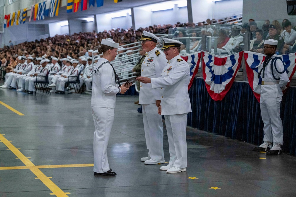 A formal U.S. Navy graduation or pass-in-review ceremony taking place in a large indoor facility, likely a drill hall or hangar. A senior naval officer in dress whites is presenting a document or certificate to a junior enlisted sailor, while another officer stands nearby. Large crowds of sailors in uniform and civilian guests fill the bleachers in the background.
