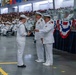 A formal U.S. Navy graduation or pass-in-review ceremony taking place in a large indoor facility, likely a drill hall or hangar. A senior naval officer in dress whites is presenting a document or certificate to a junior enlisted sailor, while another officer stands nearby. Large crowds of sailors in uniform and civilian guests fill the bleachers in the background.