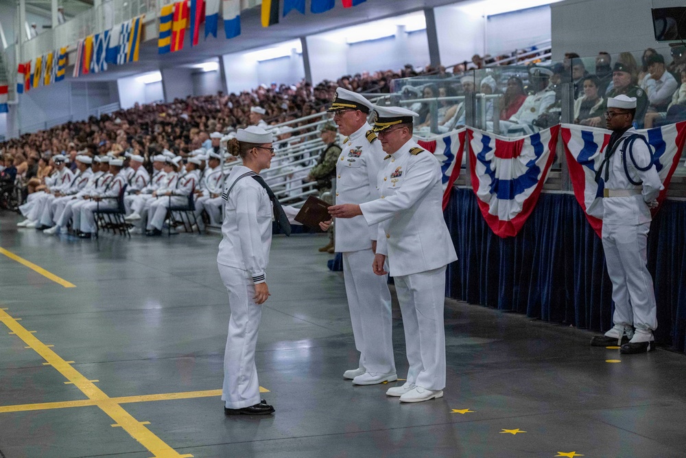 A formal U.S. Navy graduation or awards ceremony taking place in a large indoor venue, likely a drill hall or hangar. A junior enlisted sailor in dress whites is receiving an item (possibly a certificate or award) from a senior officer, also in dress whites, while another officer stands nearby. The venue is filled with hundreds of seated sailors and spectators in the bleachers, decorated with patriotic red, white, and blue bunting and nautical signal flags.