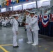 A formal U.S. Navy graduation or awards ceremony taking place in a large indoor venue, likely a drill hall or hangar. A junior enlisted sailor in dress whites is receiving an item (possibly a certificate or award) from a senior officer, also in dress whites, while another officer stands nearby. The venue is filled with hundreds of seated sailors and spectators in the bleachers, decorated with patriotic red, white, and blue bunting and nautical signal flags.