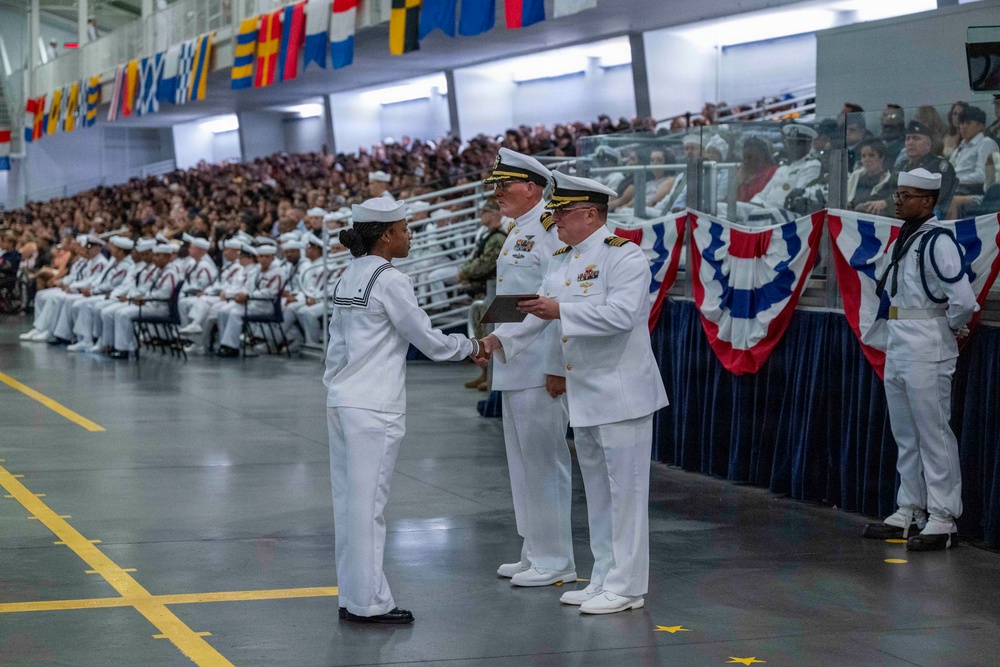 A U.S. Navy graduation or pass-in-review ceremony taking place in a large indoor hangar or drill hall. A junior enlisted sailor in dress whites is receiving a certificate or award from a senior officer, also in dress whites, while another officer stands nearby. The venue is filled with seated sailors in formation and a large civilian audience in the stands.