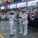 A U.S. Navy graduation or pass-in-review ceremony taking place in a large indoor hangar or drill hall. A junior enlisted sailor in dress whites is receiving a certificate or award from a senior officer, also in dress whites, while another officer stands nearby. The venue is filled with seated sailors in formation and a large civilian audience in the stands.