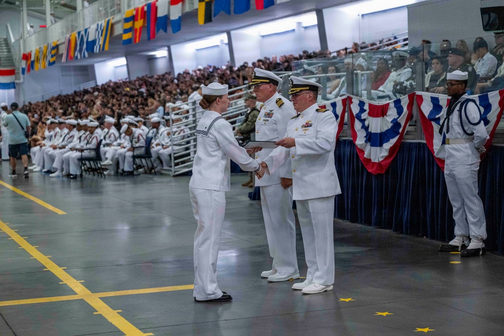 A U.S. Navy graduation or awards ceremony taking place in a large indoor facility, likely a naval training center. A female sailor in dress whites is receiving a certificate or document from a senior officer, also in dress whites, while another officer stands nearby. The venue is filled with rows of seated sailors and civilian spectators in bleachers.