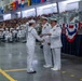 A U.S. Navy graduation or awards ceremony taking place in a large indoor facility, likely a naval training center. A female sailor in dress whites is receiving a certificate or document from a senior officer, also in dress whites, while another officer stands nearby. The venue is filled with rows of seated sailors and civilian spectators in bleachers.