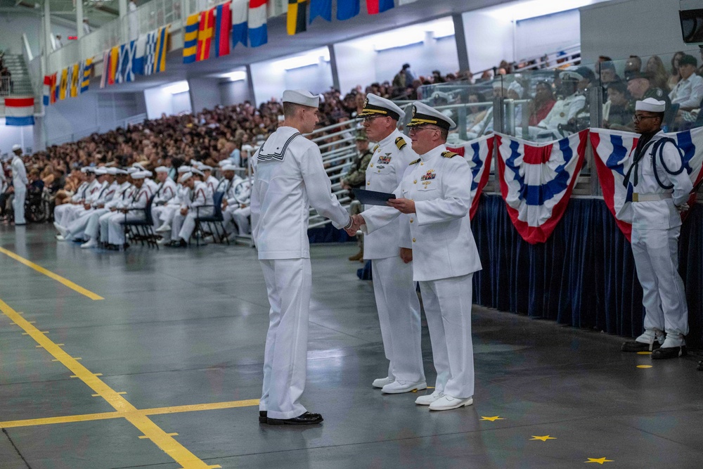 A formal U.S. Navy military ceremony is taking place in a large indoor venue, likely a hangar or gymnasium. A senior naval officer in dress whites is presenting a document or award to an enlisted sailor while another officer stands witness. A large audience of sailors and civilians observes from the background, with nautical signal flags and patriotic bunting decorating the space.