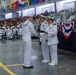 A formal U.S. Navy military ceremony is taking place in a large indoor venue, likely a hangar or gymnasium. A senior naval officer in dress whites is presenting a document or award to an enlisted sailor while another officer stands witness. A large audience of sailors and civilians observes from the background, with nautical signal flags and patriotic bunting decorating the space.