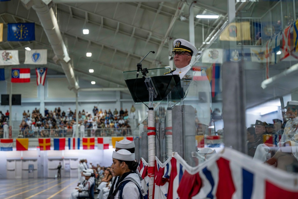 A U.S. Navy officer in dress white uniform delivers a speech at a podium during what appears to be a formal military ceremony, likely a graduation or commissioning event. The indoor venue, possibly a large gymnasium or drill hall, is decorated with numerous state and international flags hanging from the ceiling and walls. An audience including uniformed naval personnel and civilians fills the bleachers in the background.