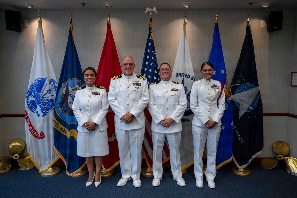 Four U.S. Navy personnel in white dress uniforms are posed for a formal group photograph in front of a display of military service flags. The setting appears to be an official military facility, likely during a ceremony or formal occasion. Two individuals appear to be senior officers based on their rank insignia and ribbons, while two appear to be junior personnel.