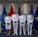 Four U.S. Navy personnel in white dress uniforms are posed for a formal group photograph in front of a display of military service flags. The setting appears to be an official military facility, likely during a ceremony or formal occasion. Two individuals appear to be senior officers based on their rank insignia and ribbons, while two appear to be junior personnel.